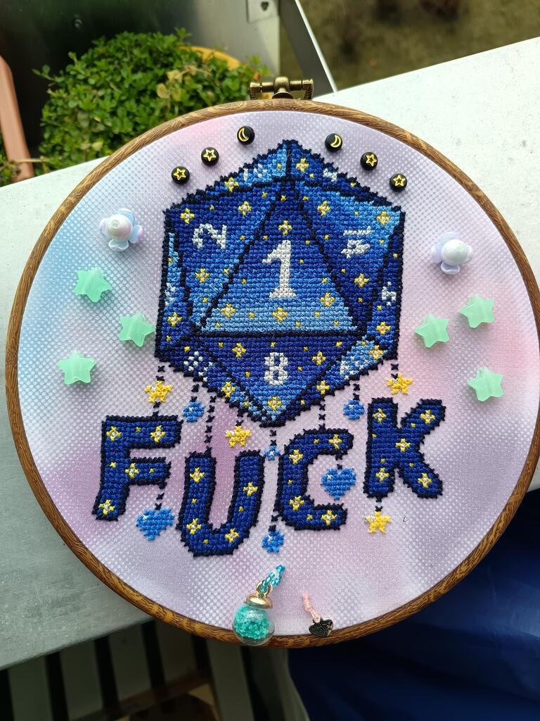 Cross stitch on pastel colored blue and pink fabric in a round hoop. The cross stitch shows a D20 with a nat1. Under it the word FUCK is written in big letters. Letters and dice are in a blue gradient with yellow stars. The numbers are white. Around the <br>cross stitch are beads and stars sewn onto the fabric. Cross stitch on pastel colored blue and pink fabric in a round hoop. The cross stitch shows a D20 with a nat1. Under it the word FUCK is written in big letters. Letters and dice are in a blue gradient with yellow stars. The numbers are white. Around the <br>cross stitch are beads and stars sewn onto the fabric.
